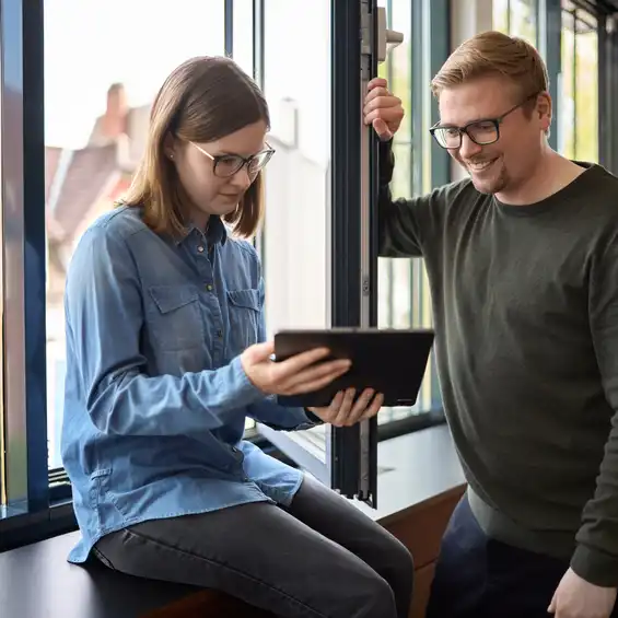 Eine Frau sitzt mit einem Tablet am offenen Fenster, neben ihr steht ein Mann