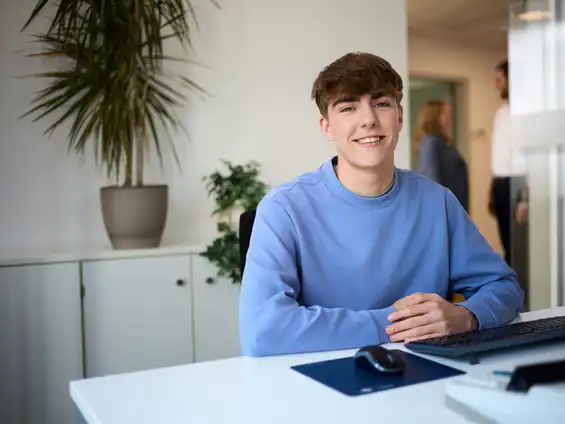 Ein Mann im blauen Sweatshirt sitzt an einem Schreibtisch und lächelt